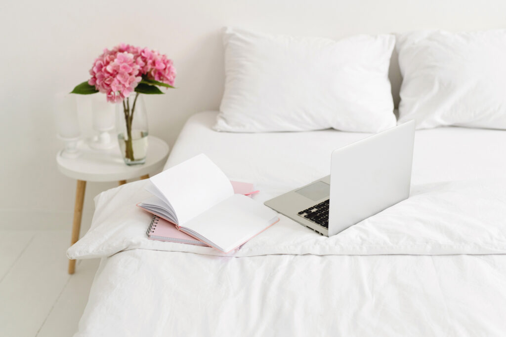 a white bedroom with a laptop and notebooks open on the bed.