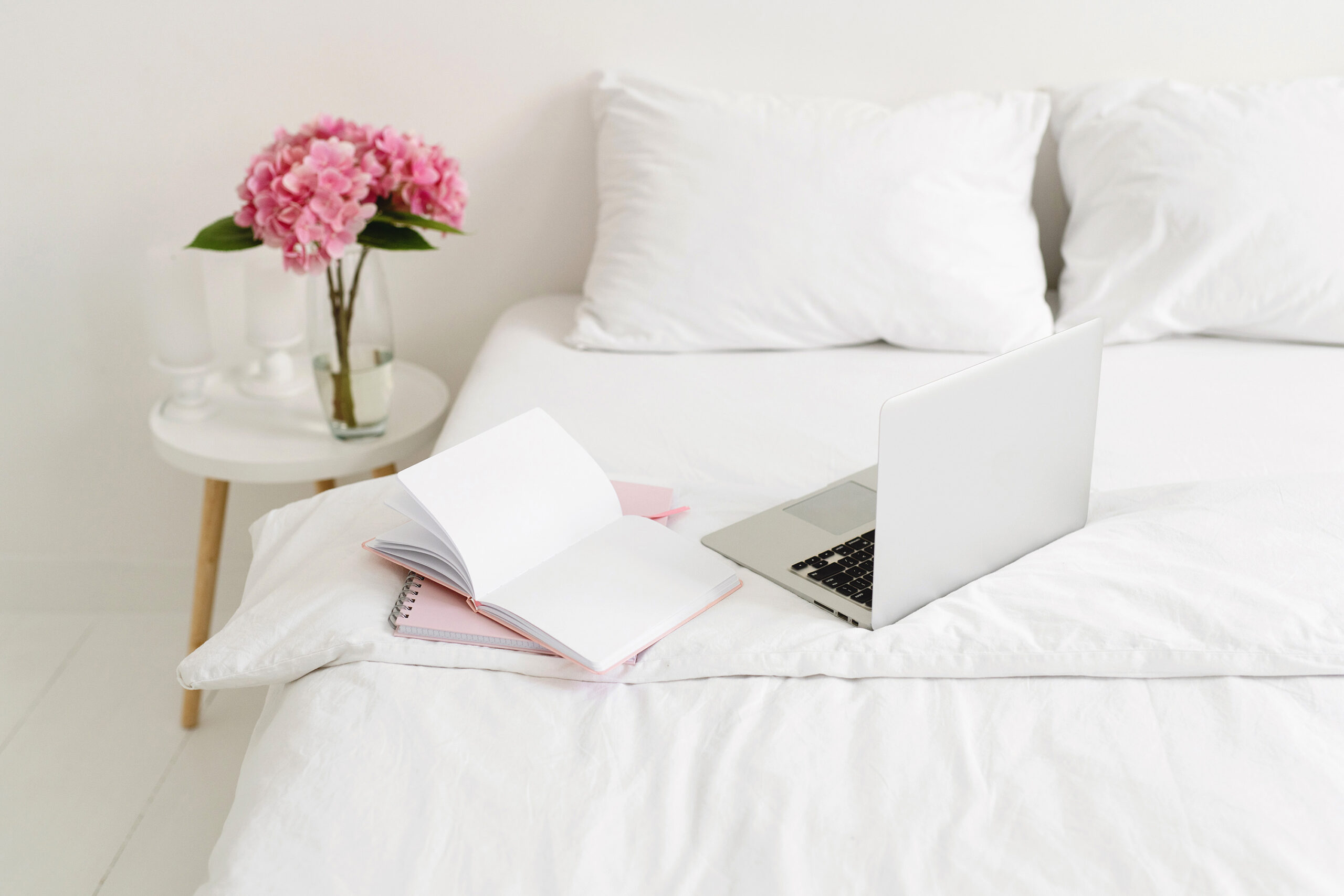 stock photo of laptop and notebooks on a white bed with pink peonies