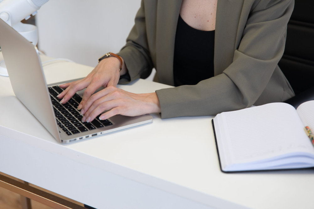 Close-up of hands typing on a laptop at a clean desk with an open notebook nearby. The professional setup suggests focus, organization, and consistent marketing efforts through writing, planning, and digital work.